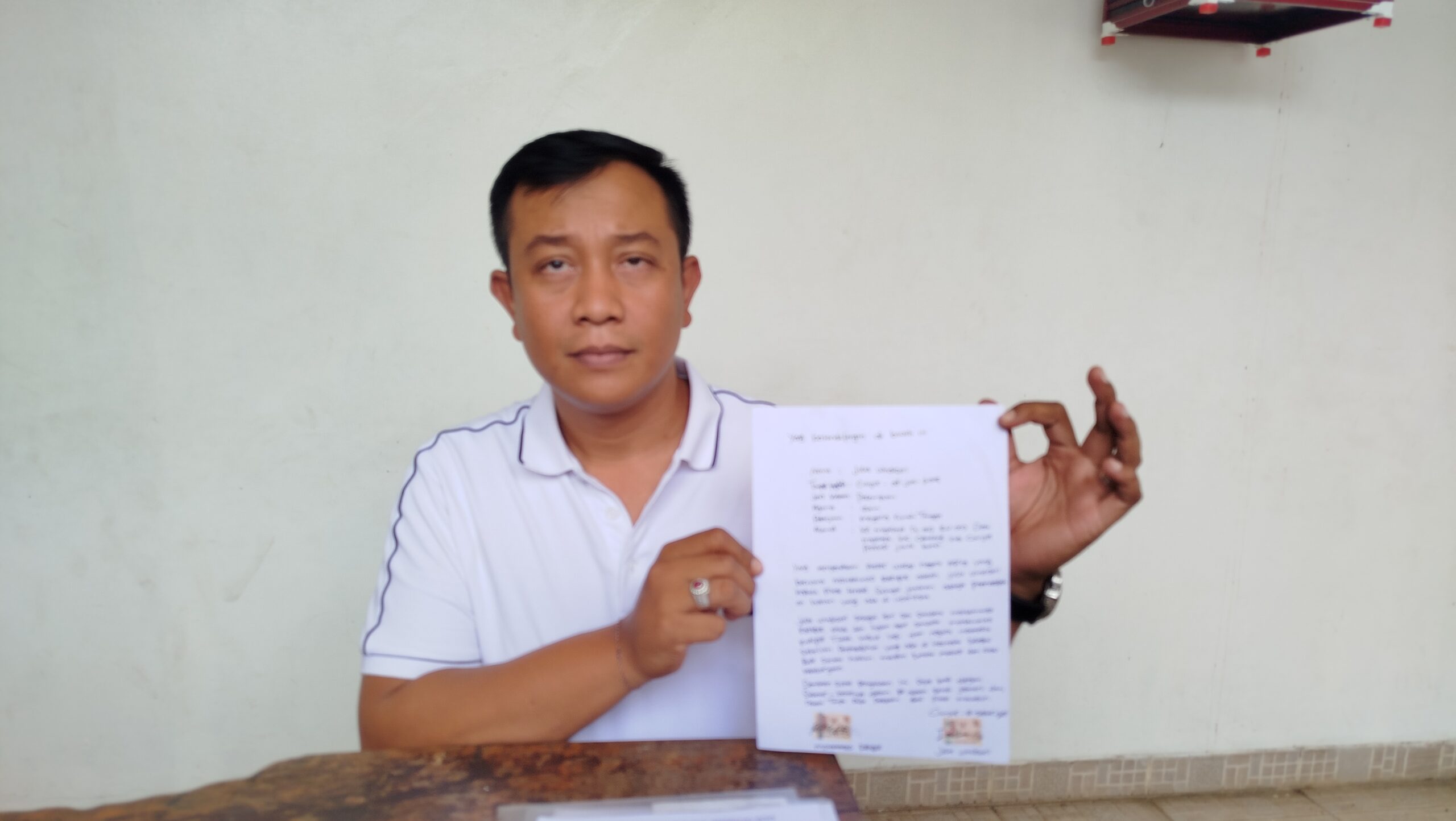 Teks: Seorang kuasa hukum mengenakan kaos putih sedang memegang dokumen laporan polisi di depannya.