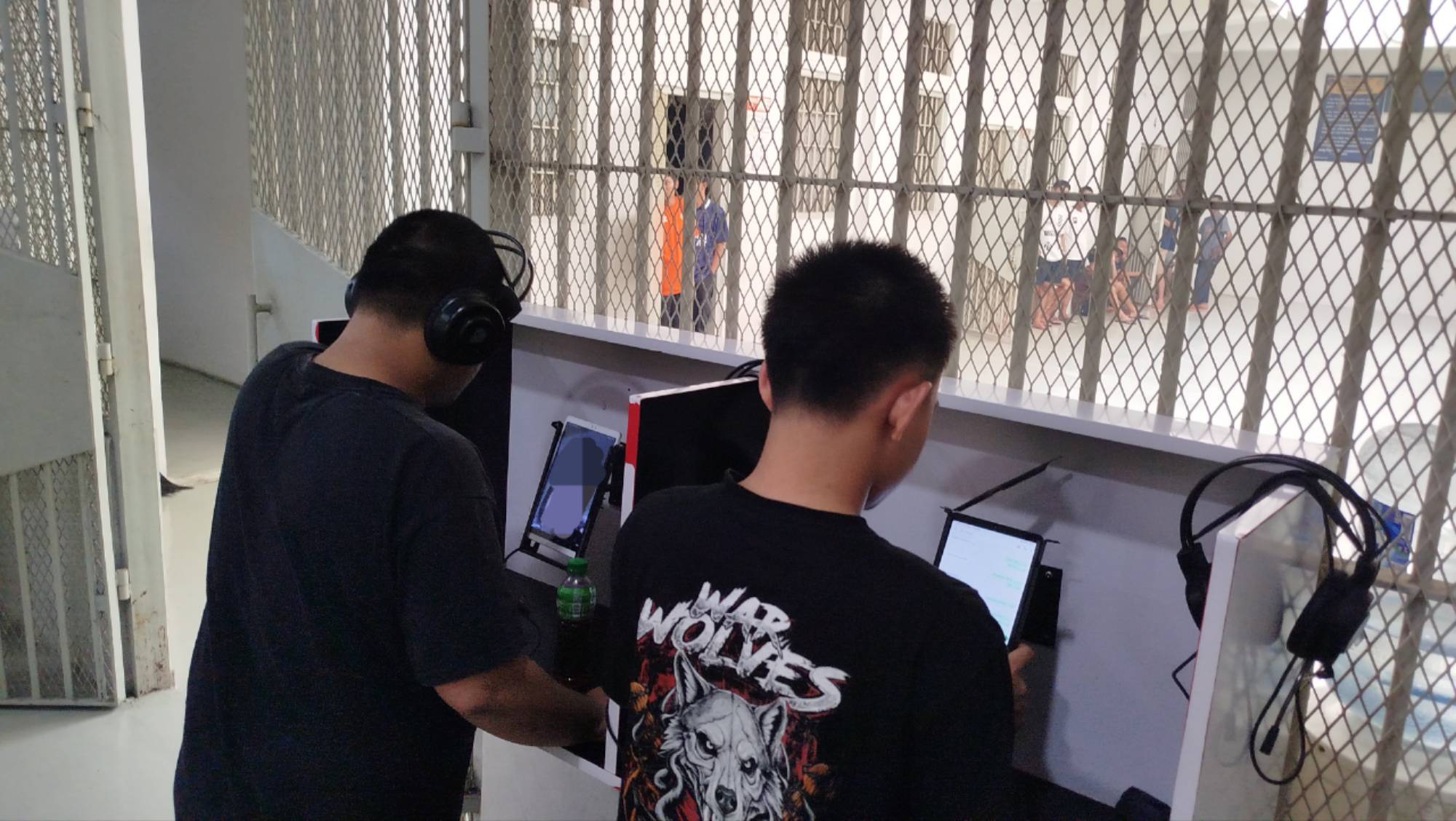 Teks: Dua warga binaan Lapas Cianjur terlihat menggunakan perangkat Wartelsus untuk melakukan panggilan suara yang diawasi, dengan area blok hunian terlihat di belakang jeruji.