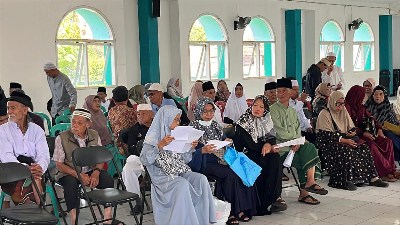 Teks: Sejumlah jemaah haji Cianjur duduk menunggu antrean vaksinasi di dalam aula asrama haji.